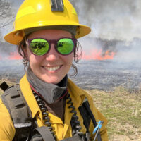 Danielle Howard stands in front of a field on fire from a prescribed burn.